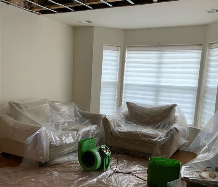 Living room with SERVPRO drying equipment by sofa and chair covered in plastic on the hardwood floor